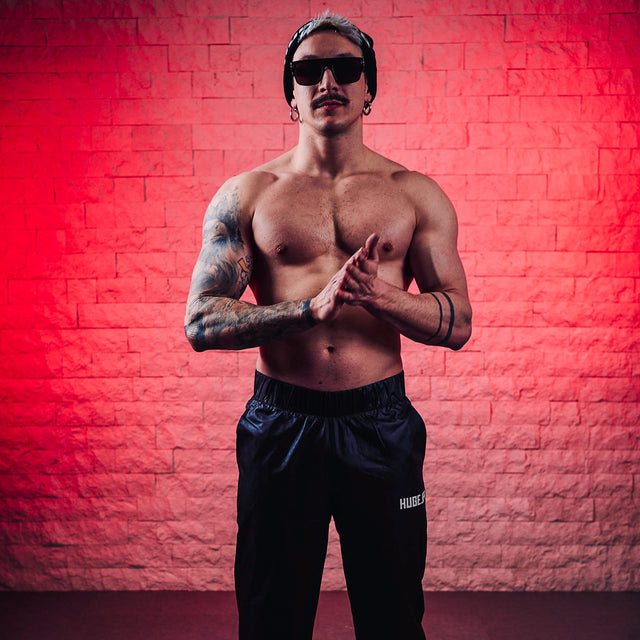 L’immagine mostra un uomo in posa con dei jogger ovvero pantaloni sportivi neri con il logo “HUGE JPG”. Lo sfondo è una parete di mattoni illuminata di rosso, che mette in risalto la figura atletica e tatuata dell’uomo, aggiungendo intensità e grinta alla composizione.