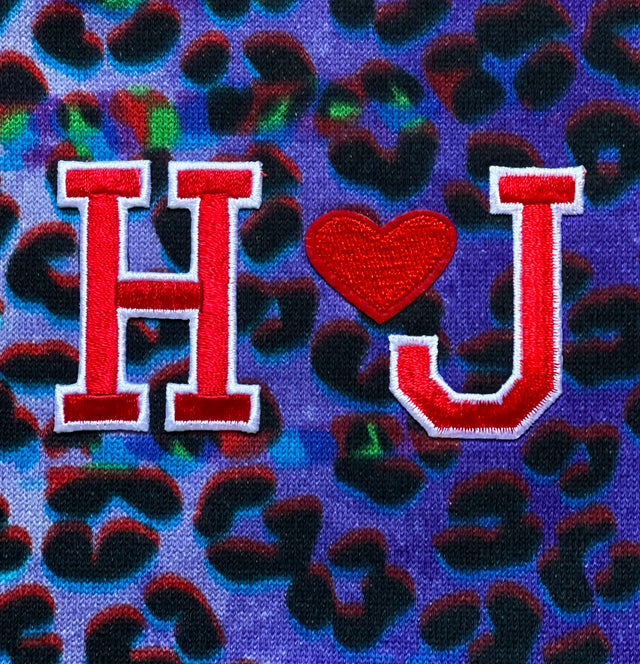 Logo ‘HUGE JPG’ ricamato sulla shopping bag ‘Sportea’ leopardata su sfondo viola. Le lettere ‘H’ e ‘J’ e un cuore, tutti ricamati, formano il logo distintivo che aggiunge un tocco personale alla borsa, esaltando il suo design unico e l’attenzione al dettaglio.