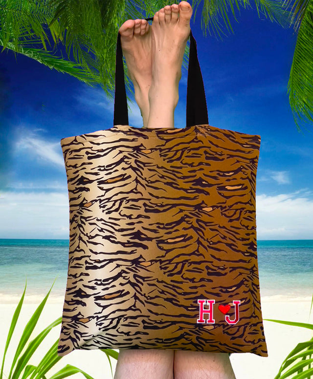 Due gambe capovolte che tengono una grande borsa da shopping SPORTEA con stampa tigrata. La borsa è posata sulla sabbia chiara di una spiaggia caraibica, con alberi tropicali ai lati dell’immagine e un mare turchese in lontananza. L’immagine evoca una sensazione di relax estivo e la borsa si presenta come un accessorio quotidiano per tutte le età e generi.