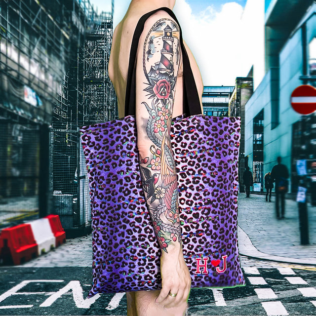 L’immagine mostra una borsa tote leopardata in tonalità di viola con le iniziali “HJ” che sta per “HUGE JPG”. La borsa è tenuta da una persona con un tatuaggio esteso sul braccio, in una strada urbana dall’atmosfera vivace, con edifici in costruzione e passanti sullo sfondo. Questo accessorio audace rappresenta un’espressione di stile individuale e moderno.